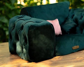 May include: A dark green velvet pet sofa with a buttoned design. It includes a small pink cushion and a dark green cushion. The sofa is on a wooden surface, with a blurred green backdrop.