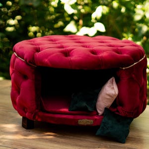 May include: A burgundy pet bed with a buttoned, rounded design and a hollow interior. The bed has a dark interior and is furnished with small cushions in pink and dark green. It rests on short, dark wooden legs.