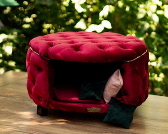 May include: A burgundy pet bed with a buttoned, rounded design and a hollow interior. The bed has a dark interior and is furnished with small cushions in pink and dark green. It rests on short, dark wooden legs.