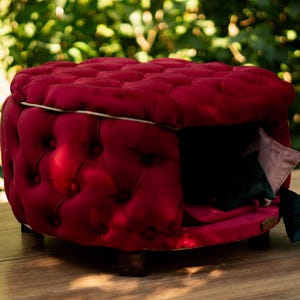 May include: A luxurious, red tufted pet bed, square in shape, with a small entrance. The bed has a dark interior and includes a small pillow. It rests on wooden legs and is placed on a wooden surface.
