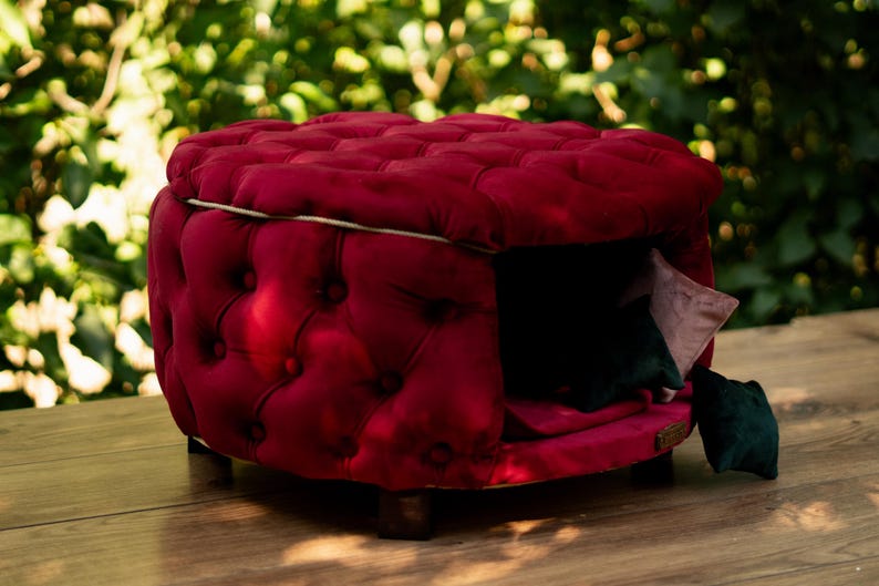 May include: A luxurious, red tufted pet bed, square in shape, with a small entrance. The bed has a dark interior and includes a small pillow. It rests on wooden legs and is placed on a wooden surface.