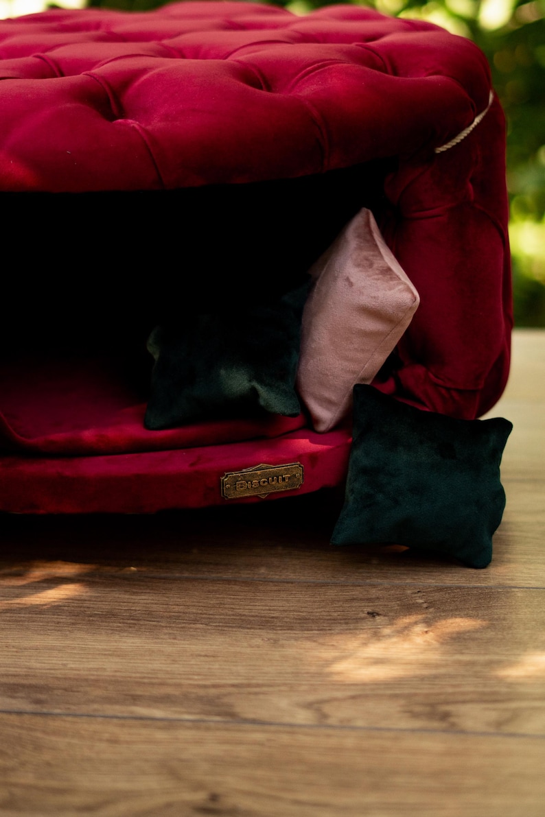 May include: A plush, claret-coloured velvet pet bed with a buttoned design. The bed features three small cushions in shades of green and pink. A gold-toned plaque with the word "Biscuit" is attached.