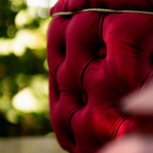 May include: Close-up of a section of a deep red velvet chair. The chair features a tufted design with buttons and a decorative rope trim. The background is blurred with green and yellow tones.