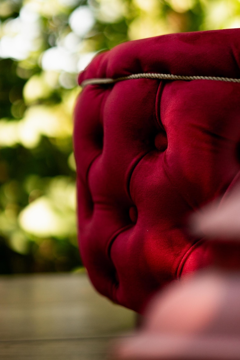 May include: Close-up of a section of a deep red velvet chair. The chair features a tufted design with buttons and a decorative rope trim. The background is blurred with green and yellow tones.