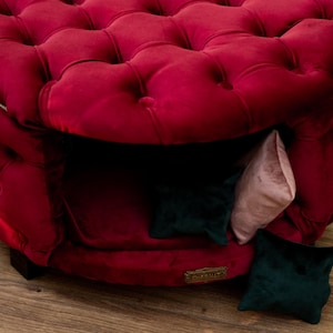 May include: A plush, burgundy velvet pet bed with a buttoned design. The bed has a cosy interior with a dark opening and includes small, decorative cushions in green and pink. A stylish and comfortable space for pets.