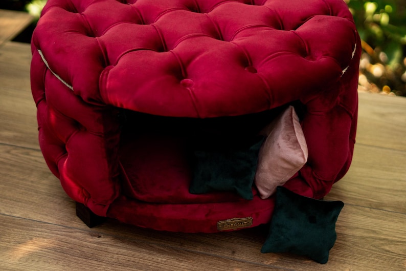May include: A plush, burgundy velvet pet bed with a buttoned design. The bed has a cosy interior with a dark opening and includes small, decorative cushions in green and pink. A stylish and comfortable space for pets.