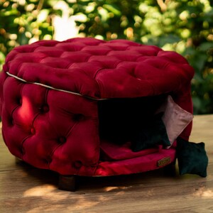 May include: A plush, burgundy pet bed with a tufted design. The bed has a square shape with a button-tufted pattern and a white trim. Inside, there are pink and green cushions. The bed is raised on small wooden feet. The bed is designed for pets.