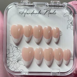 May include: A set of handmade, oval-shaped false nails in a clear plastic case. The nails have a light pink base with a white French tip. The case has the words "Handmade Nails" in cursive.