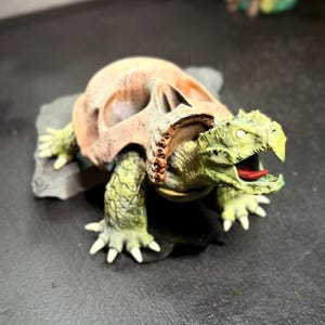 May include: A detailed sculpture of a turtle-like creature with a skull-shaped shell. The sculpture is primarily beige and green, with a red tongue visible. It is mounted on a gray stone base, showcasing intricate details.