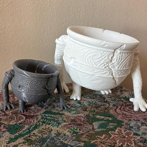 May include: Two decorative ceramic pots with legs. One is gray, the other is white. Both pots have a cracked design and are decorated with swirling patterns. The pots are standing on a patterned surface.