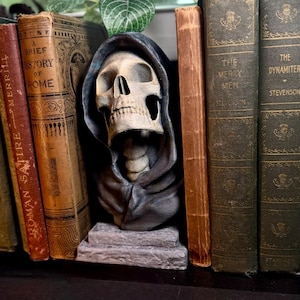 May include: A shelf display featuring antique books and gothic decor. A skull sculpture in a hooded cloak is centered among the books. The books have aged covers with titles like "Roman Satire" and "The Merry Men". Two small statues of cloaked figures flank the books.