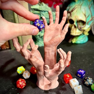 May include: A brown, hand-shaped dice holder with multiple dice scattered around it. A person's hand holds a blue and gold twenty-sided die above the holder. The dice are various colors, including red, green, and yellow, and have white or gold numbers.