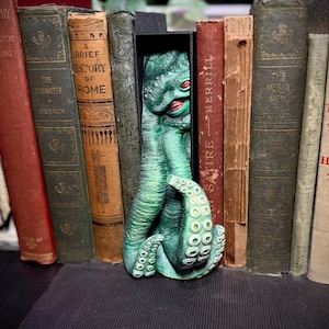 May include: A green tentacled creature bookmark with red eyes, nestled among antique books. The bookmark is detailed with suction cups and a textured surface, adding a unique touch to any reading collection. The books have titles like "A Brief History of Rome" and "The Merry Men".