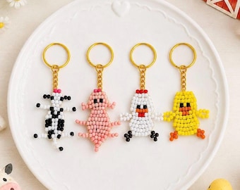 May include: Four beaded keychains featuring animal designs on a white plate. The keychains include a black and white cow, a pink pig, a white chicken with a red comb, and a yellow duck. Each charm is attached to a gold-toned metal ring.