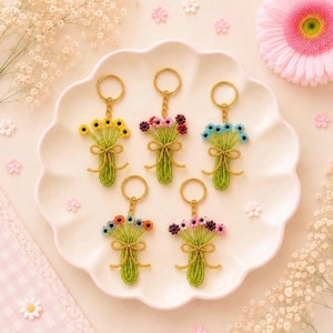 May include: Five beaded flower bouquet keychains with gold-tone rings. Each keychain features a green beaded stem bundle tied with a gold beaded bow, topped with small colorful flower heads in yellow, pink, red, blue, and multi-colored patterns.