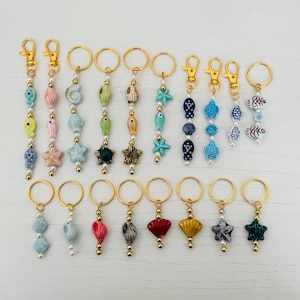May include: A collection of gold keychains with various sea-themed charms. The charms include seashells, starfish, and fish in shades of blue, green, pink, and yellow. Each keychain has a gold clasp and small pearl accents.
