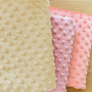 Puede incluir: Tres mantas suaves y texturizadas en tonos rosa y beige. Las mantas tienen un patrón de puntos en relieve y están dispuestas una al lado de la otra. Las mantas están hechas de un material de felpa.