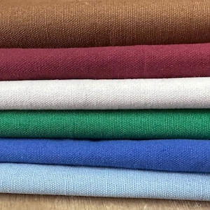 Puede incluir: Una pila de rollos de tela coloridos en tonos marrón, burdeos, blanco, verde, azul y celeste. La tela parece ser un lienzo o lino tejido apretado, adecuado para diversos proyectos de manualidades.