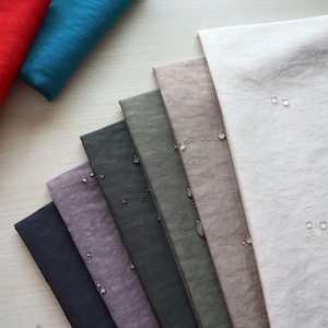 Puede incluir: Muestras de tela en varios colores, incluyendo rojo, turquesa, marino, morado y gris. La tela tiene una apariencia texturizada, y se ven gotas de agua en algunas muestras. Las muestras están dispuestas en forma de abanico.