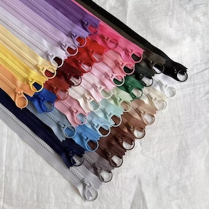 Könnte beinhalten: Eine Sammlung farbenfroher Reißverschlüsse in verschiedenen Farbtönen, darunter Lila, Rosa, Blau und Braun. Jeder Reißverschluss hat einen Metallschieber und ist diagonal vor einem weißen Hintergrund angeordnet. Ideal für Näh- und Bastelprojekte.