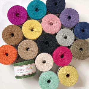 Op de afbeelding: Een verzameling kleurrijke touwspoelen in verschillende tinten, waaronder roze, teal, geel en zwart. Een groene spoel met de tekst "komorebi" is ook zichtbaar. Deze spoelen zijn ideaal voor knutsel- en decoratieprojecten.