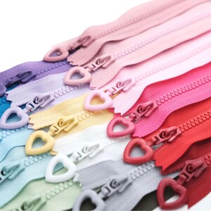 May include: A selection of colourful zippers with heart-shaped pulls. The zippers are arranged diagonally, displaying a spectrum of colours such as pink, purple, yellow, and red. The zipper teeth are visible, and the heart-shaped pulls provide a decorative element.