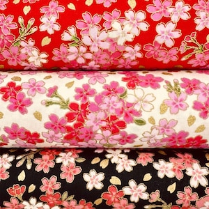 以下が含まれることがあります： 桜の花柄が繰り返される3本の布ロール。一番上のロールは赤、真ん中は白、一番下は黒です。各ロールには、花びらに金色のメタリックなアクセントが施されたピンクと白の花が描かれています。