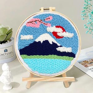 Könnte beinhalten: Ein runder Stickrahmen mit einem Berglandschaftsmotiv. Das Design zeigt einen schneebedeckten Berg, eine rote Sonne, blauen Himmel und rosa Kirschblüten. Die Stickerei wird auf einer kleinen Holzstaffelei präsentiert.