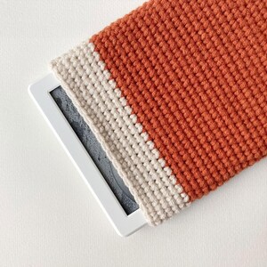 Puede incluir: Un dispositivo electrónico rectangular con marco blanco y pantalla oscura, parcialmente cubierto por una funda de ganchillo texturizada. La funda presenta una banda ancha de hilo color naranja quemado acentuada por una franja color crema.