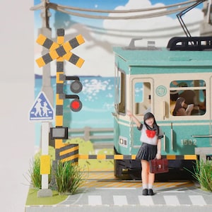 May include: A miniature paper craft diorama featuring a teal tram with the number 1502, a yellow and black railroad crossing signal, and a figure in a school uniform holding a brown bag. The background depicts a coastal scene with blue water.