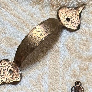Puede incluir: Un tirador de armario de color bronce con un acabado texturizado y martillado. El tirador tiene un diseño curvo con extremos decorativos en forma de corazón. El herraje es probablemente para un cajón o puerta de armario.