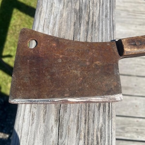 Puede incluir: Una cuchilla oxidada y vintage con mango de madera. La hoja tiene un agujero circular y se ven las palabras "COLD STEEL". La cuchilla descansa sobre tablas de madera desgastadas.