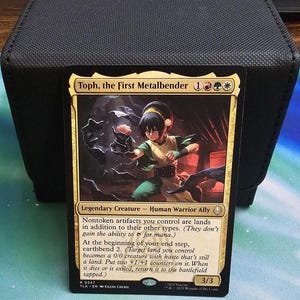 Peut inclure: Une carte à collectionner Toph, the First Metalbender représentant un personnage aux cheveux noirs vêtu de vert en position de combat. La carte comporte du texte détaillant ses capacités, une force et endurance de 3/3 et une bordure dorée.