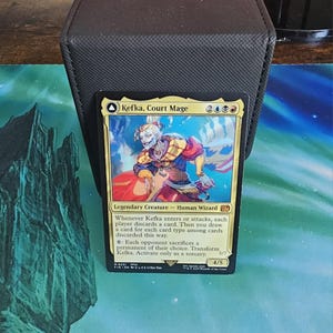 May include: A Magic: The Gathering card depicting "Kefka, Court Mage." The card shows a character in red, yellow, and blue attire. The card is set against a black card holder and a blue and green playmat. The card's text is in English.