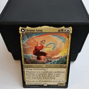 Peut inclure: Une carte à collectionner Magic: The Gathering intitulée Avatar Aang, avec une bordure dorée et une illustration d'un personnage manipulant le feu et l'eau. Le texte inclut le nom, les symboles de mana, le type de créature et 4/4.