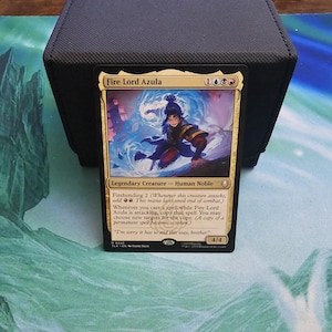 Puede incluir: Una carta de Magic: The Gathering con "Fire Lord Azula", una Criatura Legendaria. La carta tiene un borde dorado y representa a un personaje en azul y rojo con un fondo azul arremolinado. La carta está colocada contra un tarjetero negro.