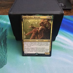 Könnte beinhalten: Eine Magic: The Gathering Sammelkarte mit dem Titel High Perfect Morcant, eine schwarze und grüne legendäre Kreaturenkarte. Das Bild zeigt einen Elfenadligen auf einem Thron. Der Kartentext enthält Fähigkeiten zu Verderben und Proliferieren.