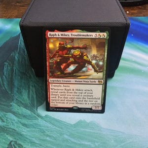 Puede incluir: Una carta de Magic: The Gathering con Raph & Mikey, Troublemakers, Tortugas Ninja Mutantes. La carta tiene un esquema de color rojo y verde con una ilustración de los personajes en acción. El texto de la carta incluye habilidades como Trample y Haste.