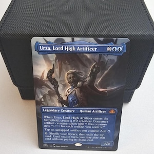 Puede incluir: Una carta coleccionable de Magic: The Gathering titulada Urza, Lord High Artificer, con una ilustración de un personaje humano artífice legendario, borde azul y texto de juego, colocada frente a una caja de almacenamiento negra texturizada.