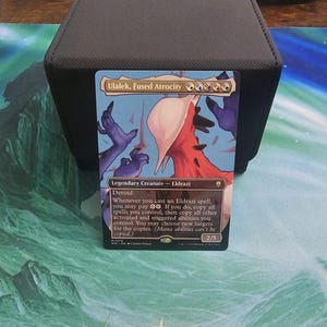Può includere: Una carta di Magic: The Gathering con "Ulalek, Fused Atrocity". La carta mostra una creatura con un design rosso e bianco su uno sfondo blu e viola. Il testo descrive le abilità della carta. La carta è in un porta carte nero.