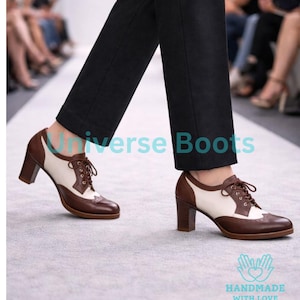 K&ouml;nnte beinhalten: Zweifarbige braune und cremefarbene Oxford-Schuhe mit Absatz und Schn&uuml;rverschluss. Die Schuhe haben einen Blockabsatz und werden mit einer schwarzen Hose getragen. Der Text "Universe Boots" ist sichtbar.