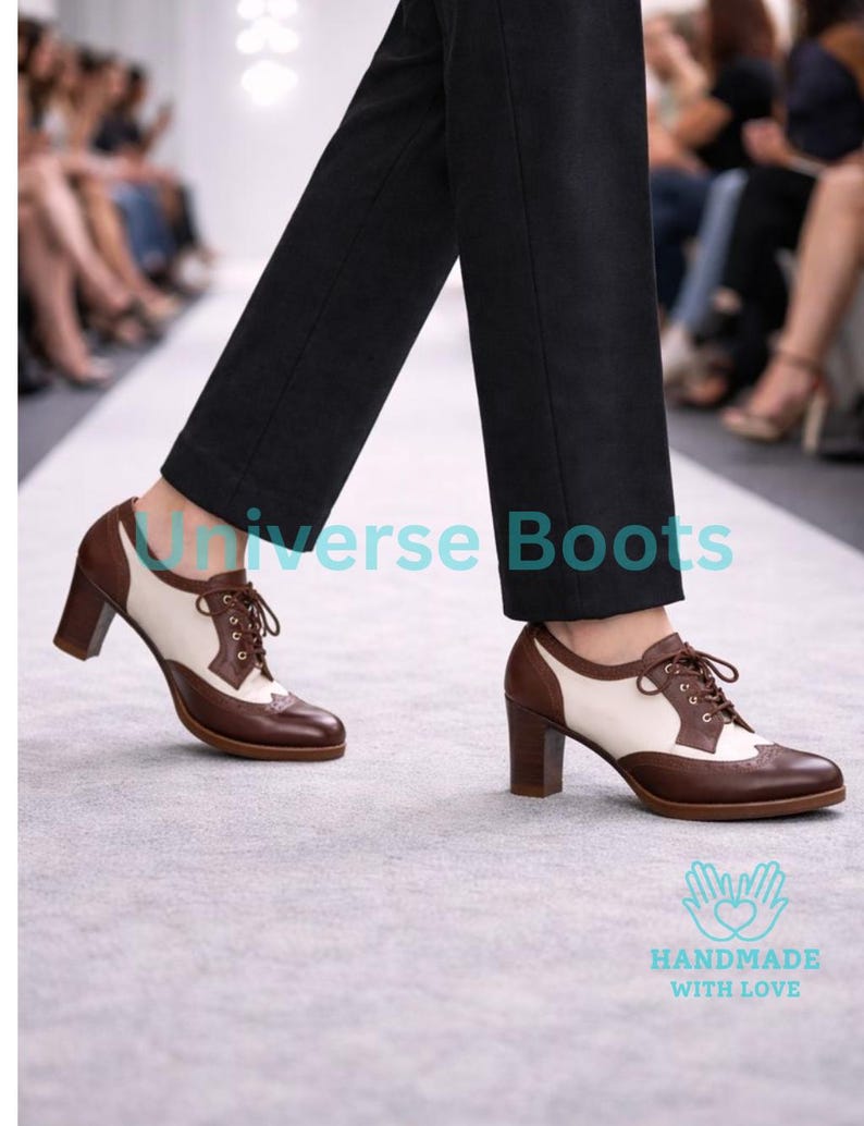 K&ouml;nnte beinhalten: Zweifarbige braune und cremefarbene Oxford-Schuhe mit Absatz und Schn&uuml;rverschluss. Die Schuhe haben einen Blockabsatz und werden mit einer schwarzen Hose getragen. Der Text "Universe Boots" ist sichtbar.