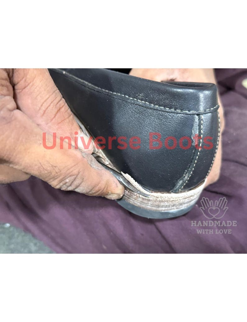 Op de afbeelding: Close-up van een zwarte leren schoen met witte stiksels en een versleten zool. De tekst "Universe Boots" is in het rood zichtbaar. De woorden "Handmade with Love" zijn ook zichtbaar.