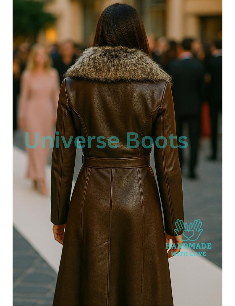 Puede incluir: Un abrigo de cuero marr&oacute;n con cuello de piel sint&eacute;tica y cintur&oacute;n en la cintura. El abrigo tiene mangas largas y una falda acampanada. El texto "Universe Boots" es visible en la parte inferior izquierda de la imagen. Tambi&eacute;n son visibles las palabras "HANDMADE WITH LOVE".