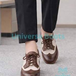 K&ouml;nnte beinhalten: Braune und cremefarbene Oxford-Schuhe mit einem klassischen Fl&uuml;gelspitzendesign. Die Schuhe haben braune Schn&uuml;rsenkel und eine robuste Sohle. Das Bild zeigt die Schuhe aus einem niedrigen Winkel, der ihre Details hervorhebt. Der Text "Universe Boots" ist sichtbar.