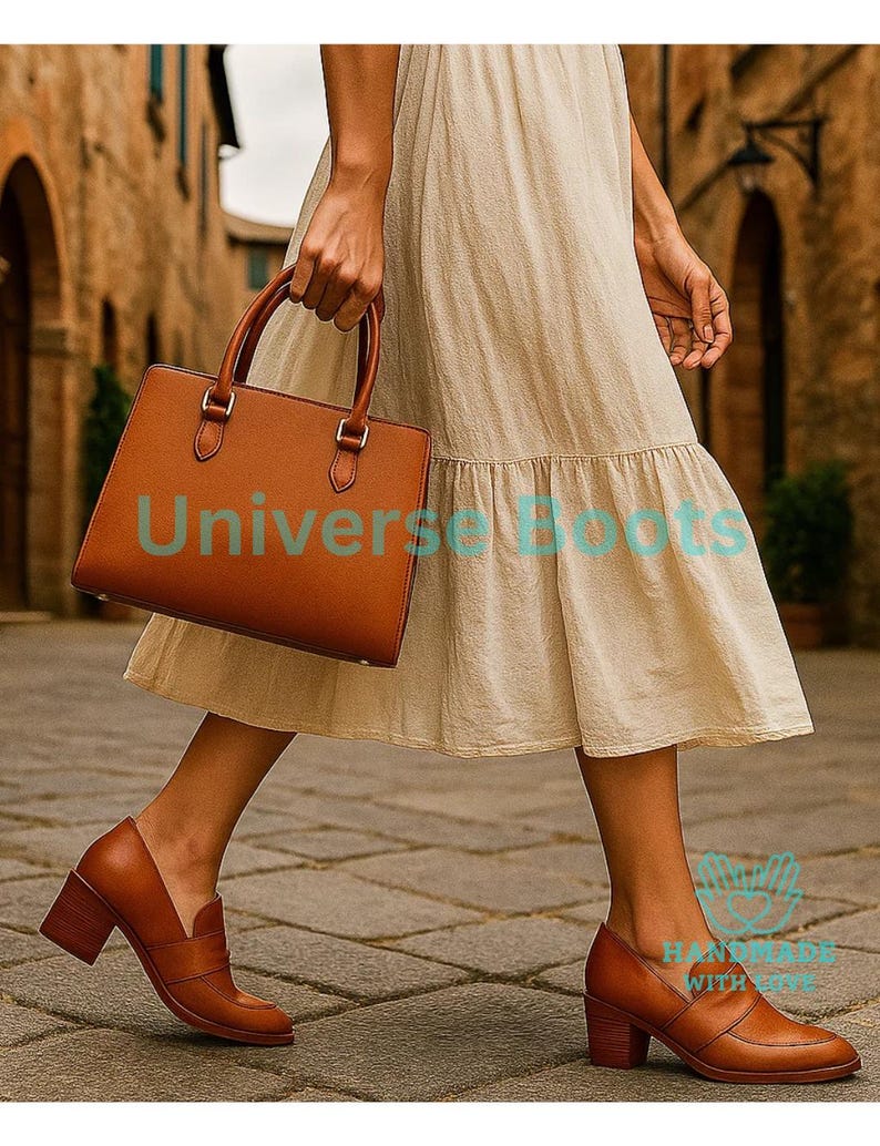 May include: A person walking in a cobbled street, wearing a beige dress and brown leather shoes. The person is carrying a brown leather handbag. The text "Universe Boots" is visible on the handbag.