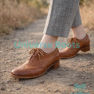 May include: Brown leather brogue shoes with a low heel and lace-up closure. The shoes have decorative perforations and are worn with gray plaid trousers. The text "Universe Boots" and "HANDMADE WITH LOVE" are visible.