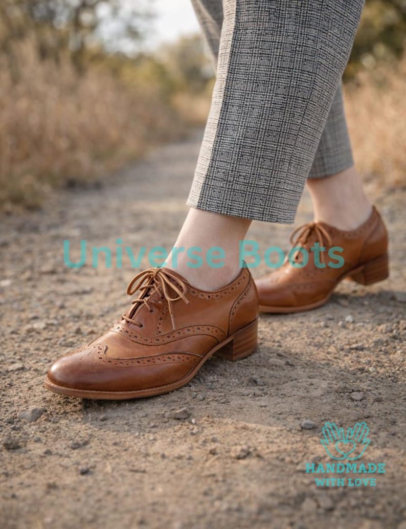 May include: Brown leather brogue shoes with a low heel and lace-up closure. The shoes have decorative perforations and are worn with gray plaid trousers. The text "Universe Boots" and "HANDMADE WITH LOVE" are visible.