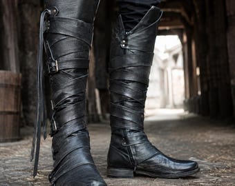 May include: A pair of black knee-high boots featuring a textured leather-like finish, multiple wrap-around straps, and side buckles. The boots have an asymmetrical top cuff, a rounded toe, and a low heel, with long leather laces hanging from the side.