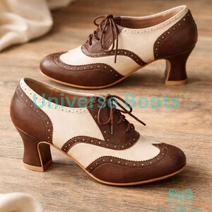 May include: A pair of two-tone brown and cream-colored oxford-style heels with a lace-up design. The shoes feature a rounded toe, a decorative perforated pattern, and a sturdy, stacked heel. The heels are approximately 2.5 inches high.